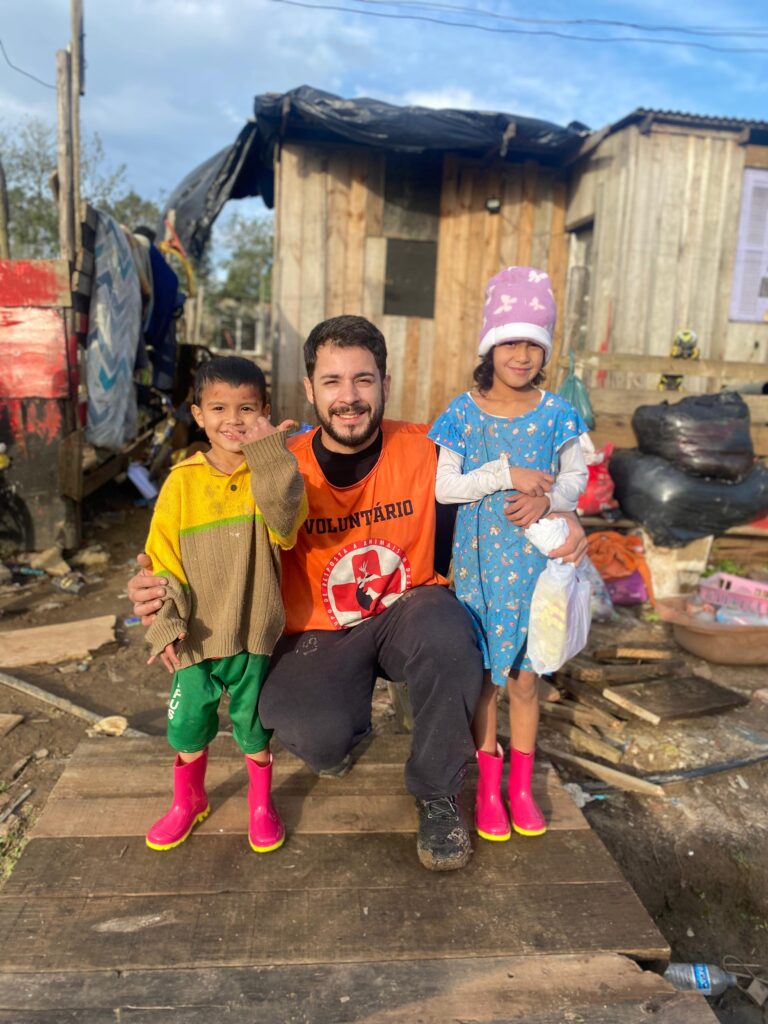 Voluntário do GRAD entrega galochas a crianças afetadas pela enchente no Rio Grande do Sul, protegendo-as da lama e dos riscos nas áreas alagadas.
